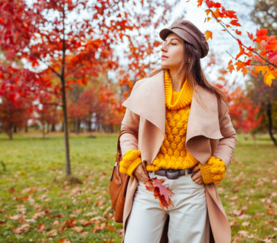 Portrait of stylish woman wears yellow sweater, coat holding handbag and red leaves walking in autumn park. Fall female clothes and accessories. Fashionable cap