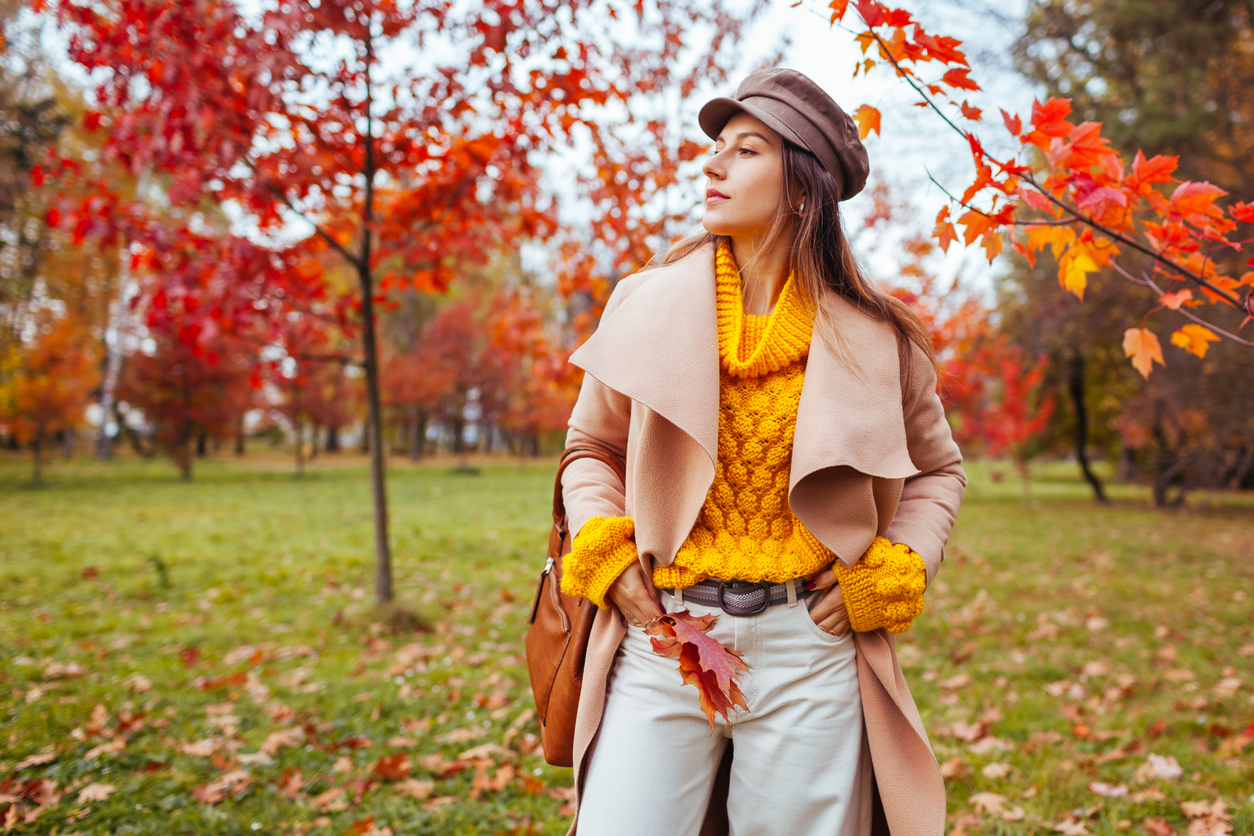 Portrait of stylish woman wears yellow sweater, coat holding handbag and red leaves walking in autumn park. Fall female clothes and accessories. Fashionable cap