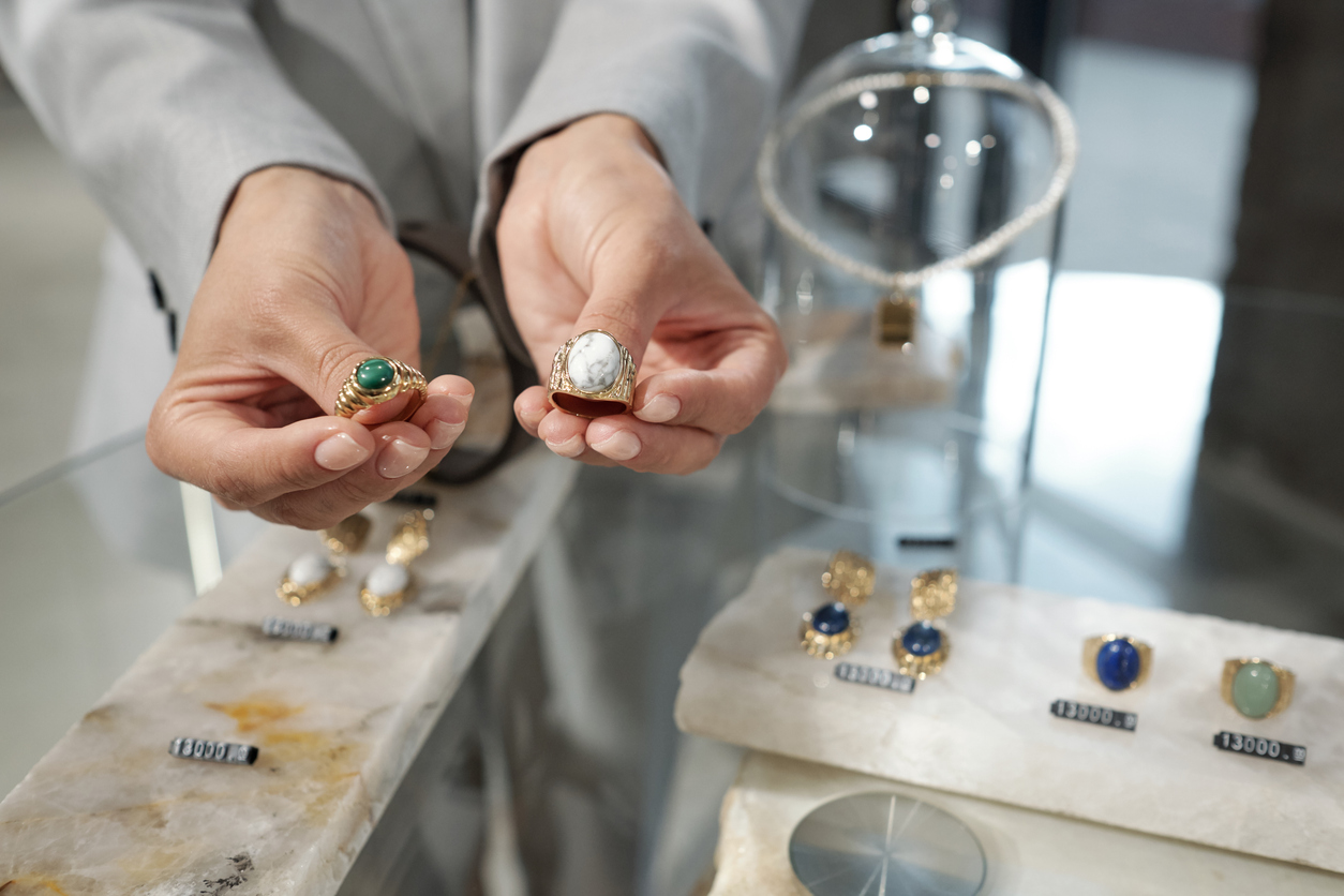 Hands of young shop assistant showing two rings to customer over display with assortment of jewelry items