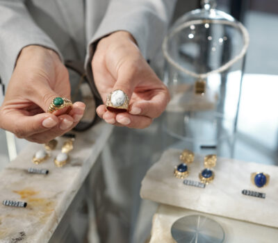 Hands of young shop assistant showing two rings to customer over display with assortment of jewelry items