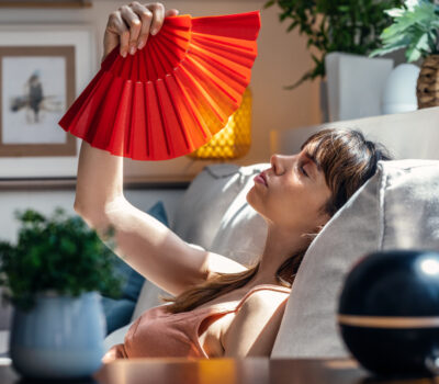 Shot of heated beautiful woman fanning herself while sitting on couch at home.